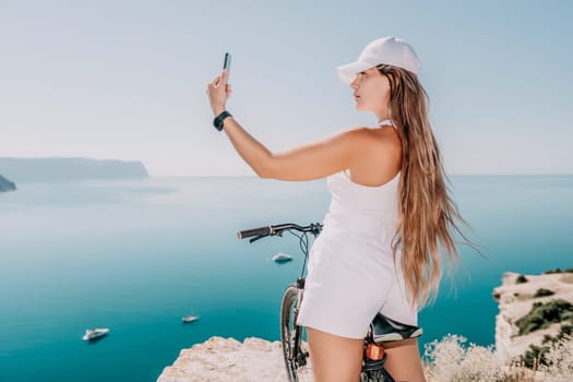 A woman cyclist on a mountain bike looking at the landscape of mountains and sea. Rear view of cyclist woman standing in front to the sea with outstretched arms. Freedom and healthy lifestyle concept