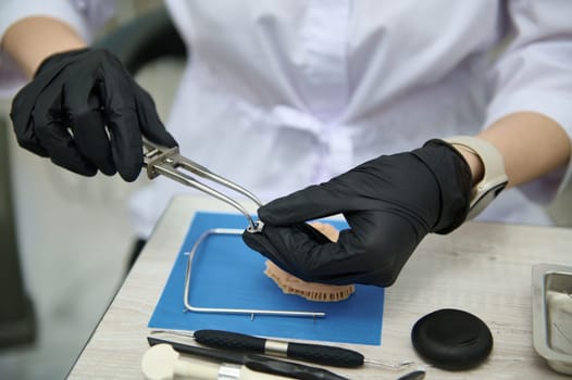 Close-up. Selective focus hands in black surgical gloves of prosthetic engineer, dental technician holding forceps, placing metal crown while working with gypsum cast of human jaw in dentistry clinic