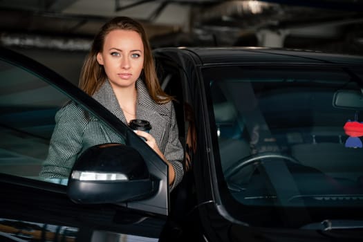 Happy woman car. she stands next to the car in the underground parking. Dressed in a gray coat, holding a glass of coffee in her hands, a black car