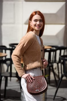 a red-haired girl in white pants and a beige sweater poses outside with a small leather handbag.
