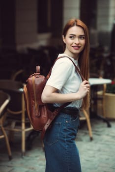 beautiful red-haired girl with red lips posing with a orange leather backpack. wearing blue jeans and a sweater