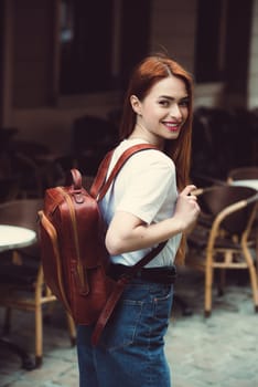 beautiful red-haired girl with red lips posing with a orange leather backpack. wearing blue jeans and a sweater