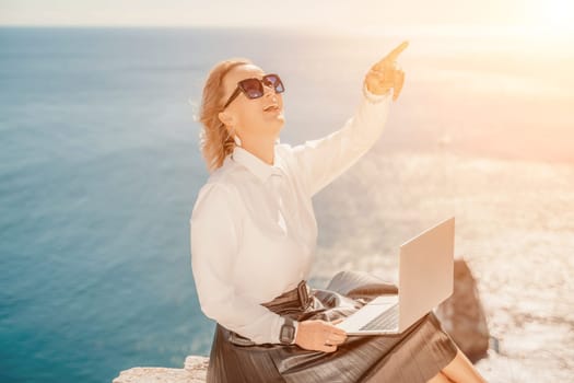 Business woman on nature in white shirt and black skirt. She works with an iPad in the open air with a beautiful view of the sea. The concept of remote work