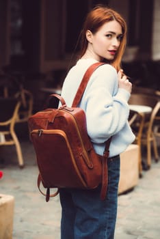 beautiful red-haired girl with red lips posing with a orange leather backpack. wearing blue jeans and a sweater