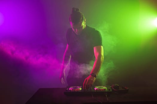 Handsome DJ in a black T-shirt at work mixing sound on her decks at a party or night club with colourful smoke light background