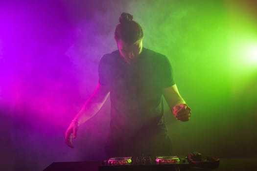 Handsome DJ in a black T-shirt at work mixing sound on her decks at a party or night club with colourful smoke light background