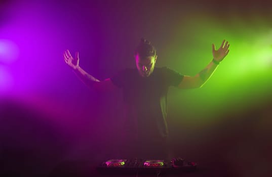 Handsome DJ in a black T-shirt at work mixing sound on her decks at a party or night club with colourful smoke light background