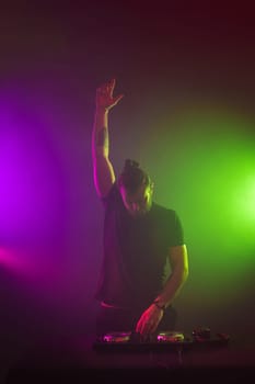 Handsome DJ in a black T-shirt at work mixing sound on her decks at a party or night club with colourful smoke light background