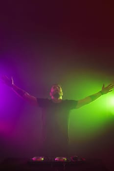 Handsome DJ in a black T-shirt at work mixing sound on her decks at a party or night club with colourful smoke light background