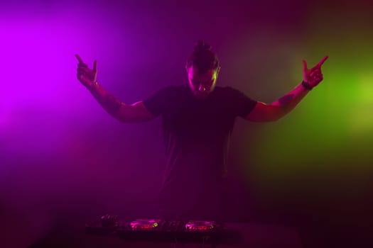 Handsome DJ in a black T-shirt at work mixing sound on her decks at a party or night club with colourful smoke light background