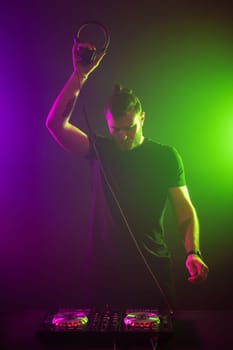 Handsome DJ in a black T-shirt at work mixing sound on her decks at a party or night club with colourful smoke light background