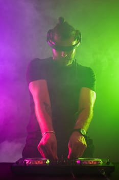 Handsome DJ in a black T-shirt at work mixing sound on her decks at a party or night club with colourful smoke light background