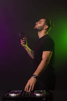 DJ playing music at mixer on colorful foggy background. A fashionable, handsome young man in a black T-shirt is behind the DJ's console. Studio shot