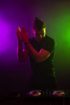 DJ playing music at mixer on colorful foggy background. A fashionable, handsome young man in a black T-shirt is behind the DJ's console. Studio shot