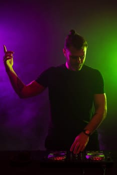 DJ playing music at mixer on colorful foggy background. A fashionable, handsome young man in a black T-shirt is behind the DJ's console. Studio shot