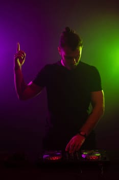 DJ playing music at mixer on colorful foggy background. A fashionable, handsome young man in a black T-shirt is behind the DJ's console. Studio shot