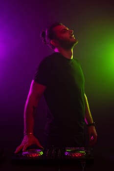 DJ playing music at mixer on colorful foggy background. A fashionable, handsome young man in a black T-shirt is behind the DJ's console. Studio shot