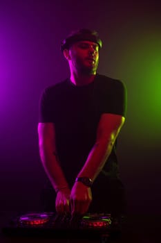 DJ playing music at mixer on colorful foggy background. A fashionable, handsome young man in a black T-shirt is behind the DJ's console. Studio shot