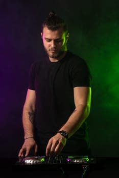 DJ playing music at mixer on colorful foggy background. A fashionable, handsome young man in a black T-shirt is behind the DJ's console. Studio shot