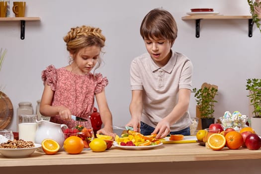 Cute cook couple. Little boy with brown hair dressed in a light t-shirt and jeans and a beautiful little girl with a braid in her hairstyle, dressed in a pink dress are cutting an orange together at a kitchen against a white wall with shelves on it.