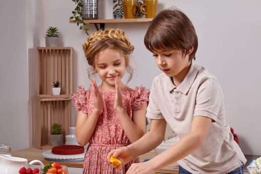 Cute cook couple. Pretty boy with brown hair dressed in a light t-shirt and jeans with a charming little girl dressed in a pink dress with a braid in her hairstyle are at a kitchen against a white wall with shelves on it. Boy is holding an orange and girl is clapping her hands.