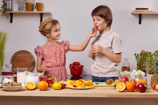Cute cook couple. Boy with brown hair dressed in a light t-shirt and jeans with a little girl dressed in a pink dress with a braid in her hairstyle are at a kitchen against a white wall with shelves on it. Girl is treating a boy with a strawberry.