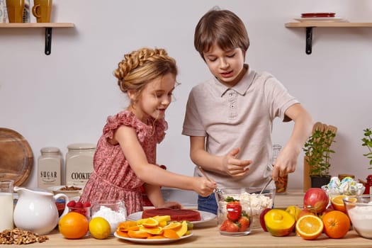 Little brunette boy dressed in a light t-shirt and jeans and a little girl with a braid in her hair, dressed in a pink dress are making a cake at a kitchen, against a white wall with shelves on it. They are taking some cottage cheese with their spoons.