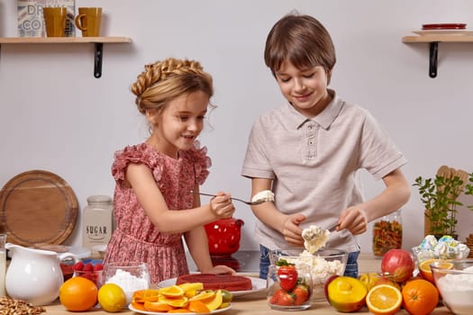 Brunette boy dressed in a light t-shirt and jeans and a pretty girl with a braid in her hair, wearing in a pink dress are making a cake at a kitchen, against a white wall with shelves on it. They are looking at a cottage cheese in their spoons.
