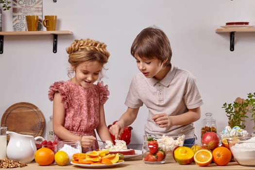 Nice boy dressed in a light t-shirt and jeans and a pretty girl with a braid in her hair, wearing in a pink dress are making a cake at a kitchen, against a white wall with shelves on it. They are spreading some cottage cheese on a cake layer.