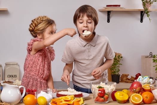 Teenage boy dressed in a light t-shirt and jeans and a beautiful girl with a braid in her hair, wearing in a pink dress are making a cake at a kitchen, against a white wall with shelves on it. Girl is treating a boy with some cottage cheese.