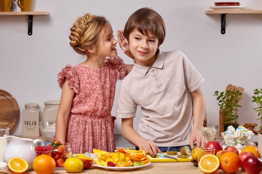 Cute cook couple. Funny boy with brown hair dressed in a light t-shirt and jeans with a wonderful little girl dressed in a pink dress with a braid in her hairstyle are at a kitchen against a white wall with shelves on it. Girl is whispering something in a boy ear.