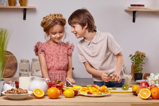 Cute cook couple. Gay boy with brown hair dressed in a light t-shirt and jeans with a little nice girl dressed in a pink dress with a braid in her hairstyle are at a kitchen against a white wall with shelves on it. Boy is cutting a kiwi and telling something to the girl.