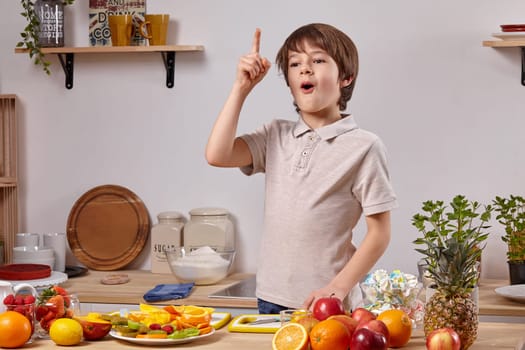 Cute little boy with brown hair dressed in a light t-shirt and jeans is cooking in a kitchen against a white wall with shelves on it. He is acting like he got an idea, put his index finger up and opened his mouth widely.