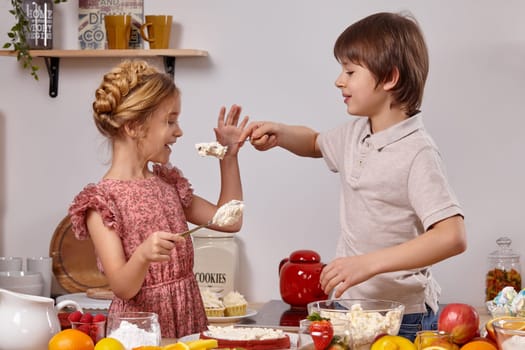 Handsome boy dressed in a light t-shirt and jeans and a beautiful girl with a braid in her hair, wearing in a pink dress are making a cake at a kitchen, against a white wall with shelves on it. Boy is treating a girl with some cottage cheese.
