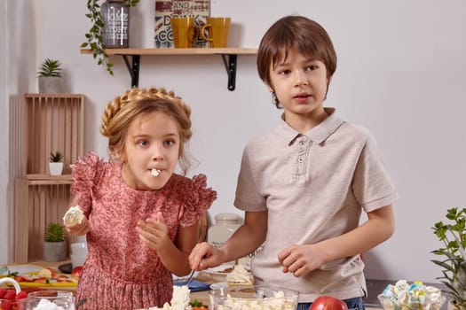 Kid dressed in a light t-shirt and jeans and a beautiful girl with a braid in her hair, wearing in a pink dress are making a cake at a kitchen, against a white wall with shelves on it. Girl is eating a cottage cheese and they a wonderly looking away.