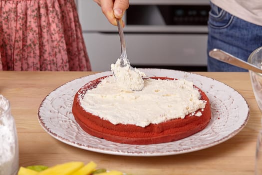 Close-up shot of a boy dressed in a light t-shirt and jeans and a beautiful girl wearing in a pink dress are making a cake at a kitchen, against a white wall with shelves on it. Boy is spreading some cottage cheese on a cake layer with a spoon.