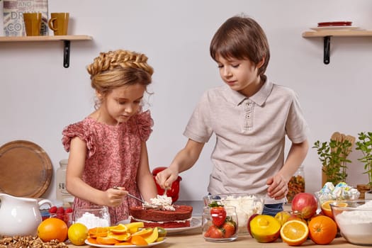 Brunette boy dressed in a light t-shirt and jeans and a cute girl with a braid in her hair, wearing in a pink dress are making a cake at a kitchen, against a white wall with shelves on it. They are spreading some cottage cheese on a cake layer.