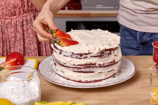 Close-up shot of a child dressed in a light t-shirt and jeans and a girl wearing in a pink dress are making a cake at a kitchen. Boy is decorating a cake with some strawberries.