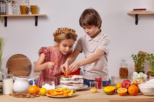 Brunette kid dressed in a light t-shirt and jeans and a beautiful girl with a braid in her hair, wearing in a pink dress are making a cake at a kitchen, against a white wall with shelves on it. They are decorating it with some strawberries.