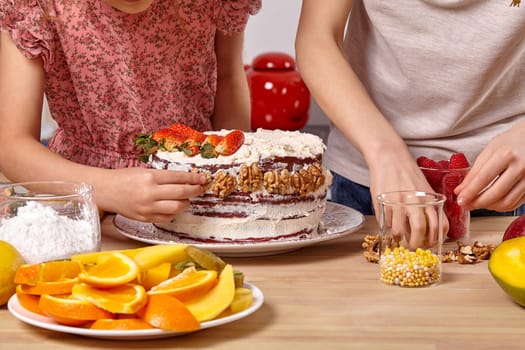 Close-up shot of a child dressed in a light t-shirt and jeans and a girl wearing in a pink dress are making a cake at a kitchen. They are decorating it with some walnuts.