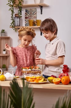 Little brunette boy dressed in a light t-shirt and jeans and a beautiful girl with a braid in her hair, wearing in a pink dress are making a cake at a kitchen, against a white wall with shelves on it. They are decorating it with some raspberries. Photo shot through a palm leaves.