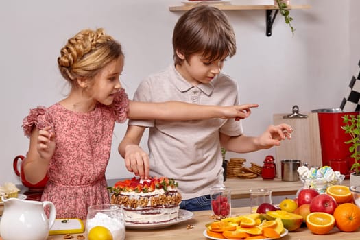 Boy dressed in a light t-shirt and jeans and a girl with a braid in her hair, wearing in a pink dress are making a cake at a kitchen, against a white wall with shelves on it. They are decorating it whith a marshmallow.