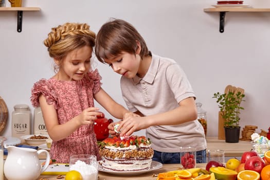 Handsome boy dressed in a light t-shirt and jeans and a beautiful girl with a braid in her hair, wearing in a pink dress are making a cake at a kitchen, against a white wall with shelves on it. They are carefully decorating it with some marshmallow.
