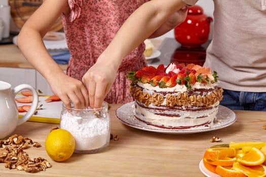 Close-up shot of a boy dressed in a light t-shirt and jeans and a girl wearing in a pink dress are making a cake at a kitchen. They are taking some powdered sugar from a jar.