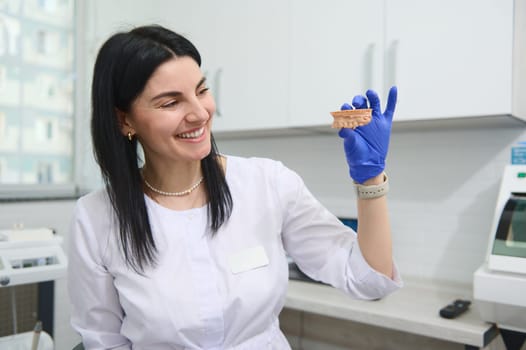 Beautiful experienced female dentist doctor, dental technician, prosthetic engineer smiling, showing at camera the gypsum mold of human upper jaw in her hands. Dental practice. Orthodontics. Dentistry