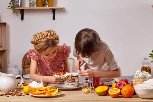 Brunette child dressed in a light t-shirt and jeans and a pretty girl with a braid in her hair, wearing in a pink dress are making a cake at a kitchen, against a white wall with shelves on it. They are decorating it with some walnuts.