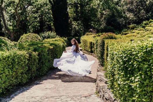 Brunette runs white dress park. A beautiful woman with long brown hair and a long white dress runs along the path along the beautiful bushes in the park, rear view.