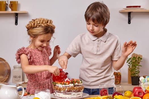 Little boy dressed in a light t-shirt and jeans and a girl with a braid in her hair, wearing in a pink dress are making a cake at a kitchen, against a white wall with shelves on it. Children sprinkle it with some powdered sugar.