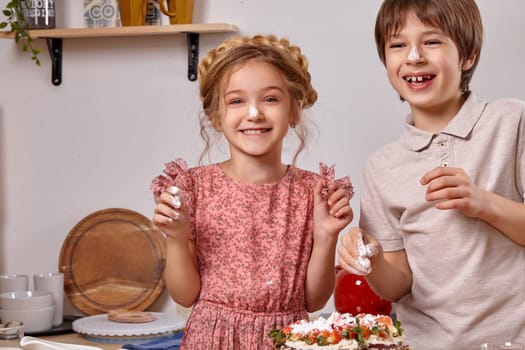Funny boy dressed in a light t-shirt and jeans and a pretty girl with a braid in her hair, wearing in a pink dress are making a cake at a kitchen, against a white wall with shelves on it. They have smeared noses to each other with an icing sugar and laughing.
