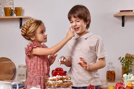 Boy dressed in a light t-shirt and jeans and a charming girl with a braid in her hair, wearing in a pink dress are making a cake at a kitchen, against a white wall with shelves on it. Girl is smearing a nose of a boy with a powdered sugar.